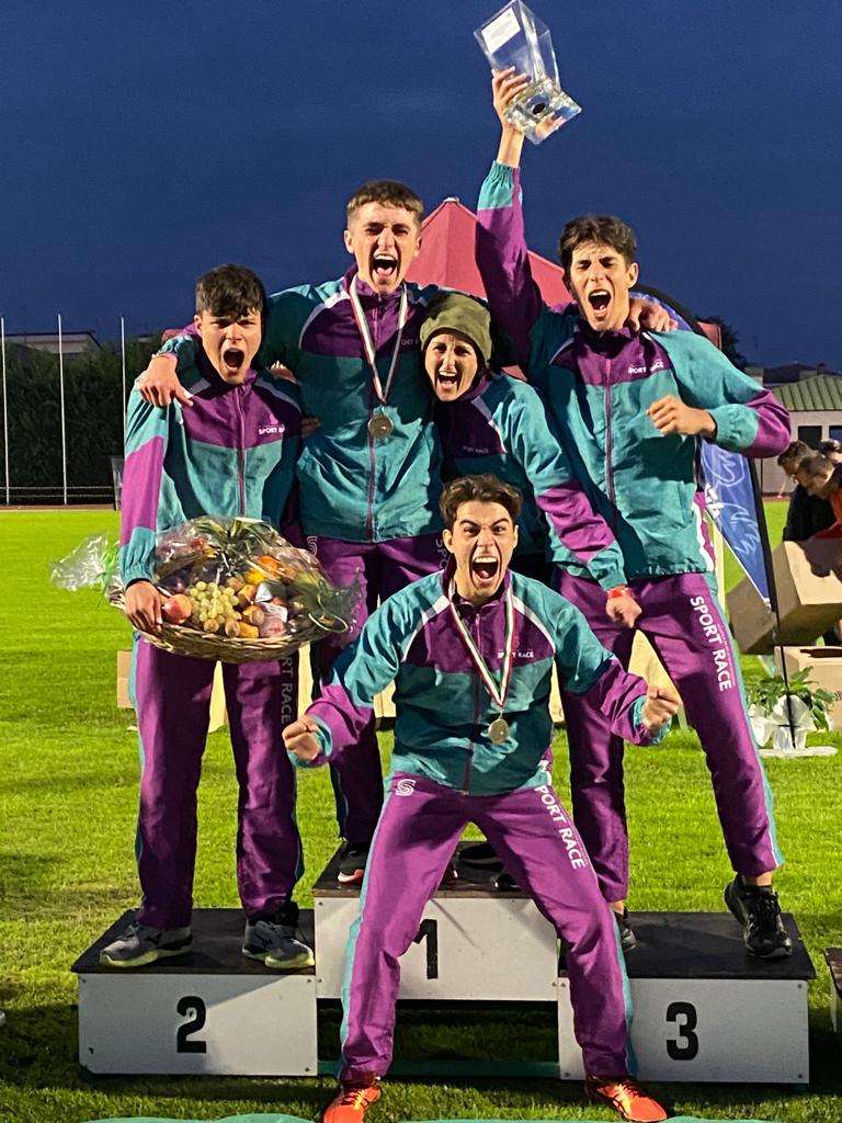 La squadra vincente della staffetta festeggia sul podio dei vincitori, tenendo in mano il trofeo e il cesto di frutta. {{brizy_dc_image_alt imageSrc=